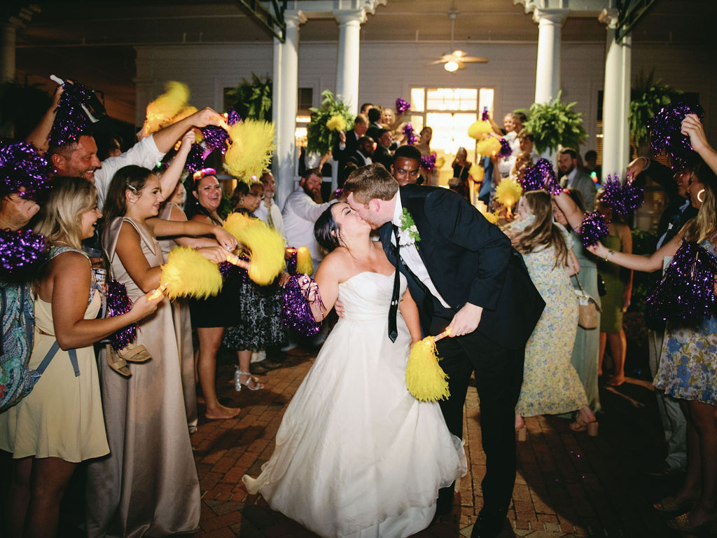 Couple exits their wedding while their guests shake purple and yellow pom poms