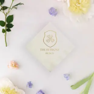 an ivory beverage napkin with a monogrammed crest foil printed on top, with a lavender background and spring pastel wedding flowers all around