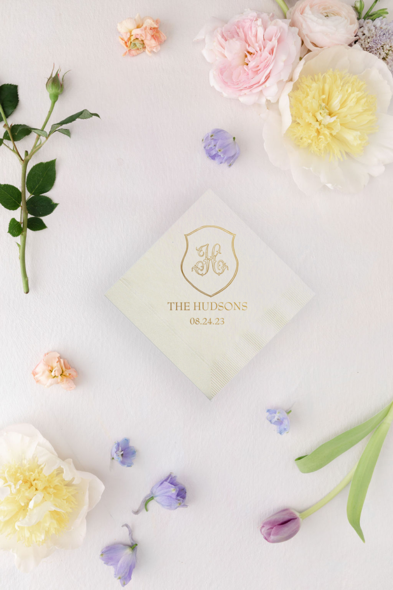 an ivory beverage napkin with a monogrammed crest foil printed on top, with a lavender background and spring pastel wedding flowers all around