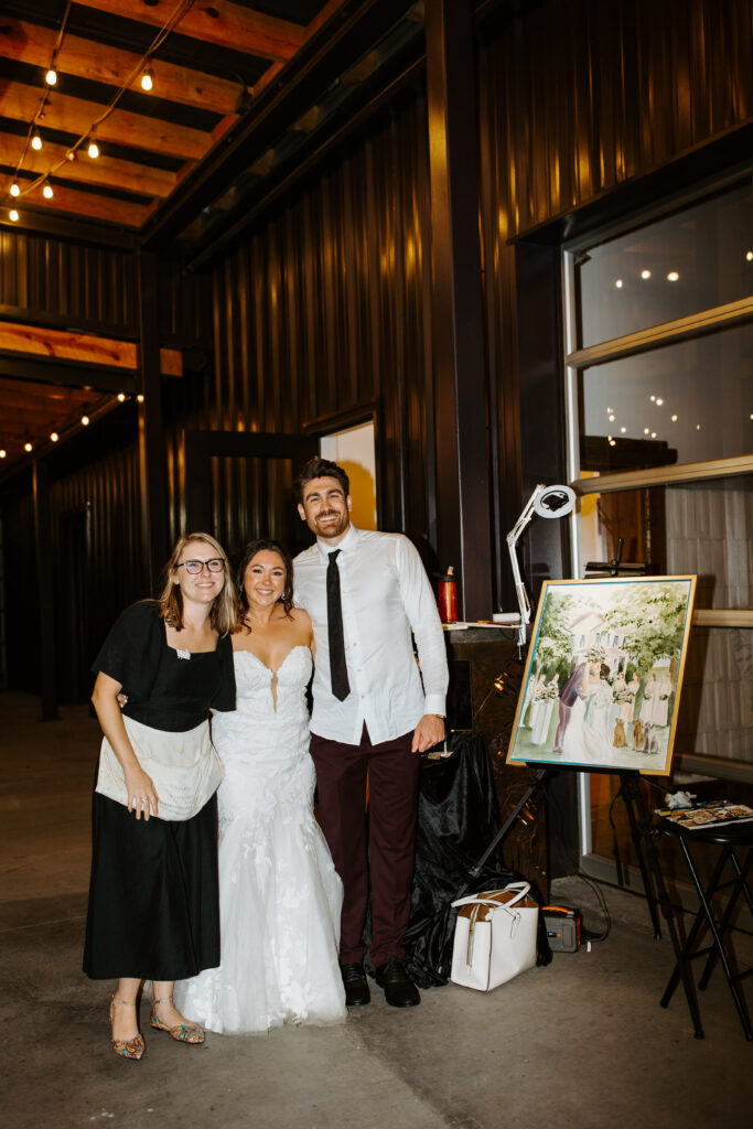 live wedding painter Ashley Triggiano and couple Sydney and hunter with their live painting portrait at the meadows Raleigh nc with a rustic barn background and market lights