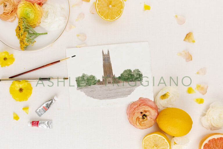 Duke Chapel watercolor painting for wedding invitations shown on a pink background with paintbrushes and citrus fruit as a flatlay photo