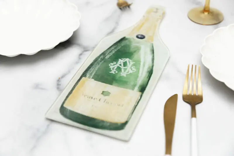 handpainted champagne bottle monogrammed gift cutting board for engagement parties on a marble background with gold cutlery