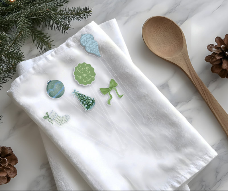 green and blue watercolor illustrations on acrylic stir sticks placed on a white tea towel with Christmas items along the border