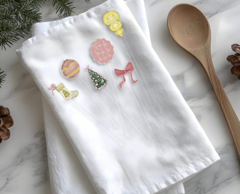 pink and green watercolor illustrations on acrylic stir sticks placed on a white tea towel with Christmas items along the border